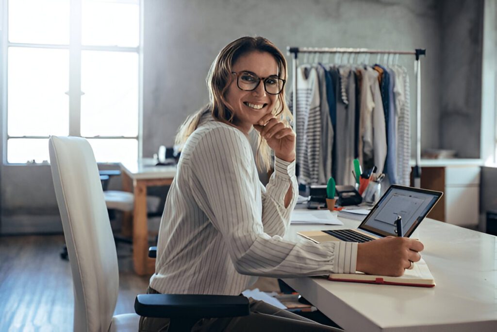 A woman in glasses and a striped shirt sits at a desk with a laptop, writing in a notebook. A rack of clothing is in the background, and sunlight filters through a window.