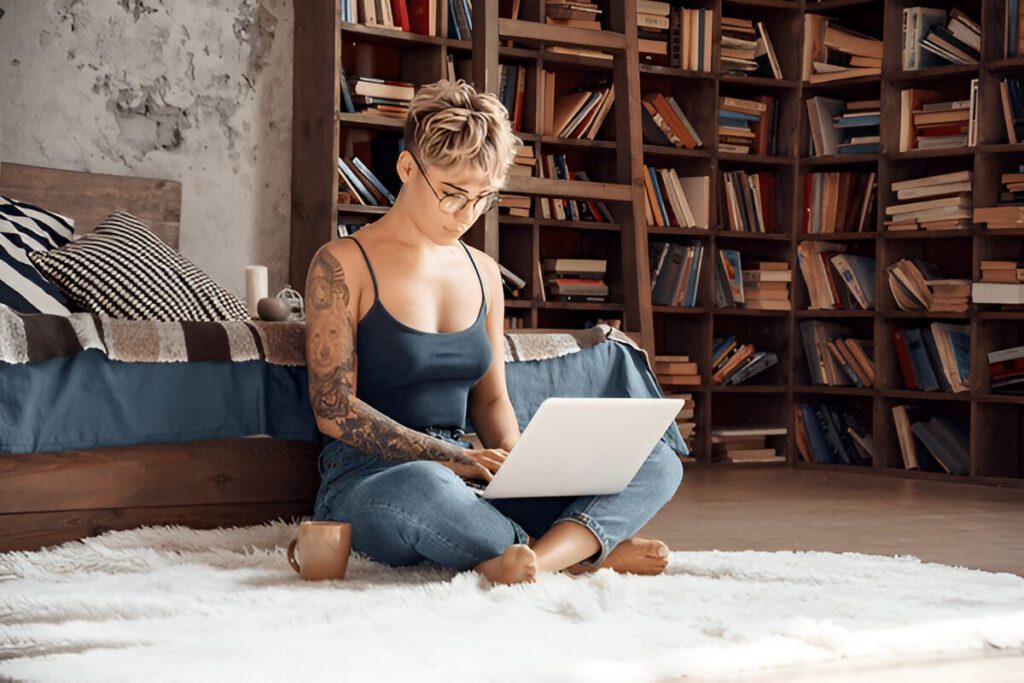 Person sitting on a rug in front of a bookcase, working on a laptop. They have short hair, glasses, and tattoos. A bed and a mug are nearby.