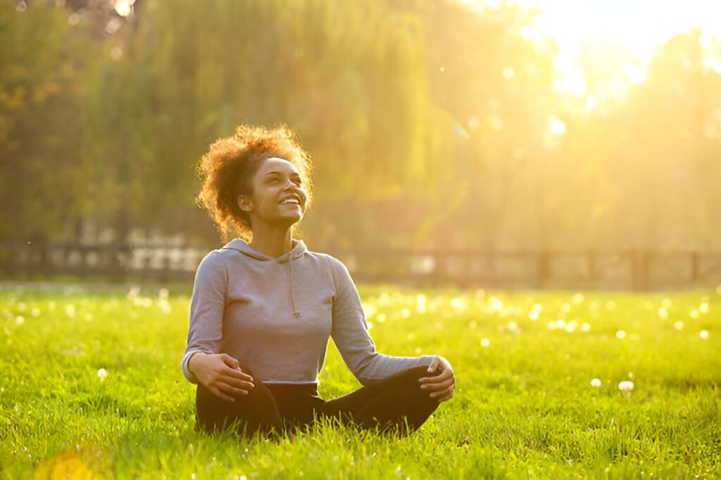 A person sits cross-legged on grassy field, smiling under bright sunlight, wearing a gray hoodie and black pants, with trees and soft sunlight in the background.