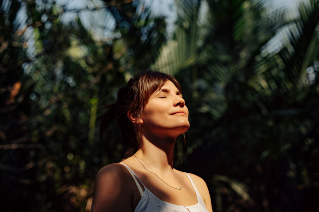 A woman in a white tank top stands in a sunlit forest with eyes closed and a serene expression.